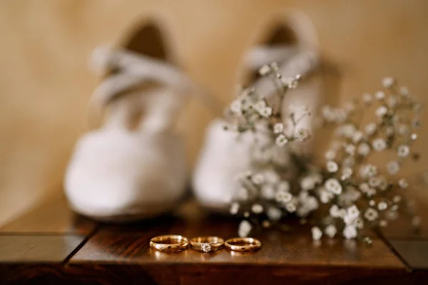 Anelli nuziali e anello di fidanzamento su un tavolo di legno con scarpe bianche da sposa e fiori di campo. — Foto Stock