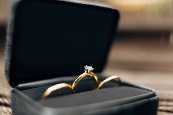 Close-up de alianças de casamento e um anel de noivado com uma grande jóia em uma caixa de veludo preto contra um fundo borrado. — Fotografia de Stock