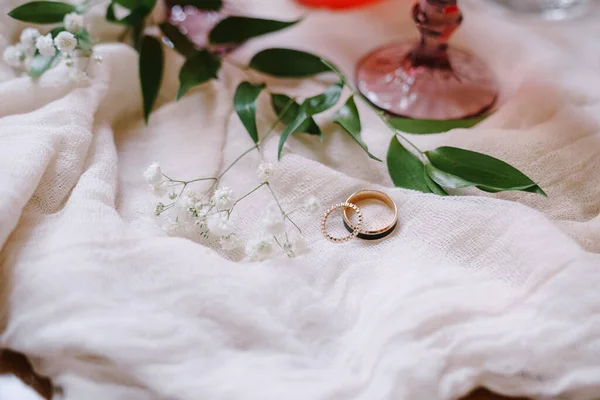 Anéis de casamento da noiva e noivo em um tecido branco com um ramo com folhas verdes e pequenas flores brancas. — Fotografia de Stock