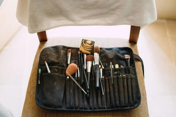 Czarna praca kosmetyczka kosmetyczna torba z pędzlami do makijażu, gąbka i pęseta brwi na drewnianym krześle. — Zdjęcie stockowe