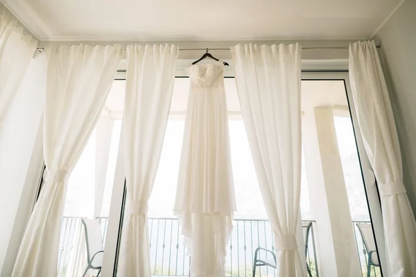 Langes weißes Kleid der Braut mit einem Spitzenkorsett auf einem schwarzen Kleiderbügel an der Decke auf dem Gesims vor dem Hintergrund weißer Vorhänge im Zimmer. — Stockfoto