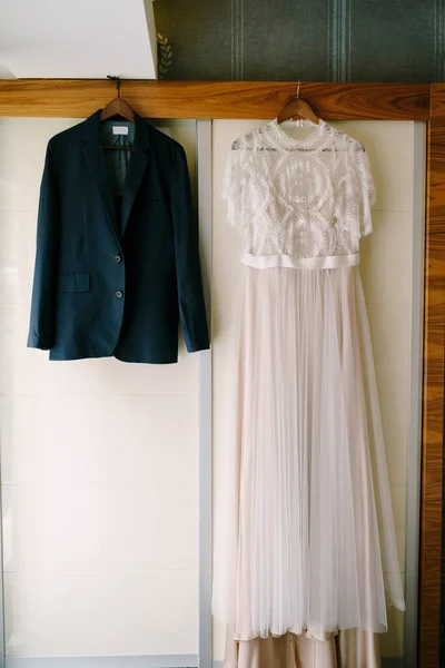 Vestido de novia blanco de la novia y chaqueta del novio en perchas de madera en la habitación contra la pared. —  Fotos de Stock