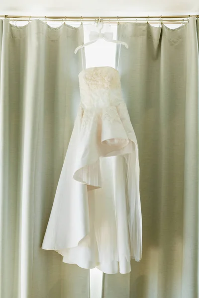 Белое платье невесты оригинального дизайна на вешалке с бабочкой жениха на фоне занавесок. — стоковое фото