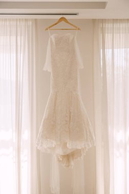 Ahşap bir askıda beyaz perdelerin arkasında beyaz bir gelinlik..