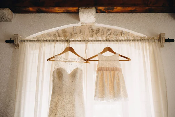 Cornice con cortina blanca y perchas de madera con vestido de dama de honor y vestido de bebé blanco. —  Fotos de Stock