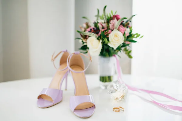 Sandalias de novia lila sobre la mesa con los anillos de novia y novio y un ramo de flores en un jarrón. —  Fotos de Stock