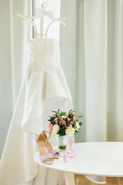 Sandalias lila sobre una mesa blanca con un ramo de flores junto a la ventana con cortinas y un vestido de novia en una percha con corbata de lazo en la habitación. —  Fotos de Stock