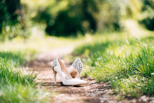 Chaussures de mariage pour femmes blanches de la mariée, à talons hauts, décorées de pierres à bijoux. Sur un sentier forestier parmi l'herbe verte. — Photo