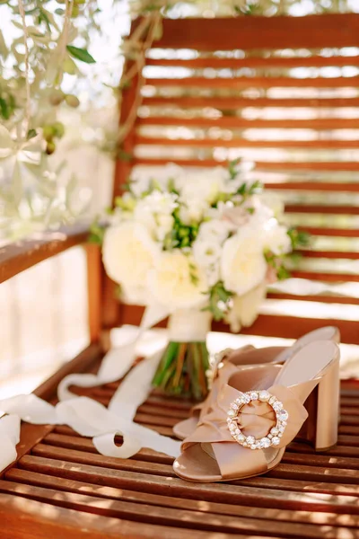 Zuecos beige con hebilla redonda en una silla de madera con un ramo de flores blancas atadas con una cinta. — Foto de Stock