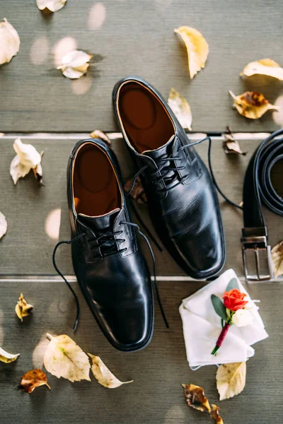 Close-up de sapatos pretos de um homem com atacadores desatados, em uma textura de madeira com um cinto e noivos boutonniere, com folhas de outono amarelas. — Fotografia de Stock