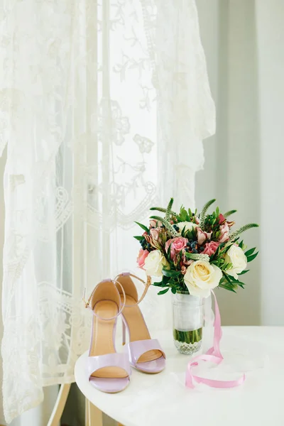 Sandalias lila sobre una mesa blanca junto a la ventana con un velo nupcial blanco y un ramo de flores en un jarrón en la habitación. —  Fotos de Stock