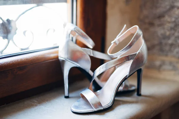 Silverfärgade damsandaler. Pärlemor sandaler av bruden på fönsterbrädan vid fönstret med en brun träram. — Stockfoto