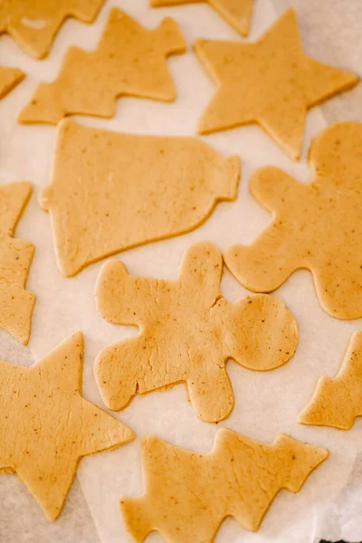 Processo de fazer biscoitos caseiros ou pão de gengibre em diferentes formas. Os biscoitos de massa crua estão em uma assadeira coberta com pergaminho para assar antes de serem enviados para o forno. Fechar — Fotografia de Stock