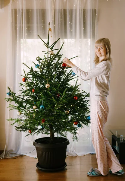 Mladá dívka v pyžamu zdobí vánoční stromek s míčky. — Stock fotografie