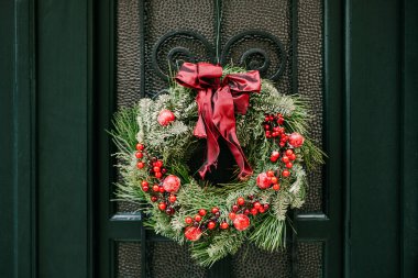 Kırmızı yay ve böğürtlenli, köknar ve çam dallarından oluşan bir Noel çelengi, sahte bir kafesle yeşil bir ahşap kapıya asılır..