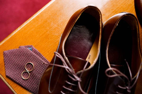 Close-up de sapatos masculinos com gravata e anéis de casamento da noiva e do noivo em uma mesa de madeira. — Fotografia de Stock
