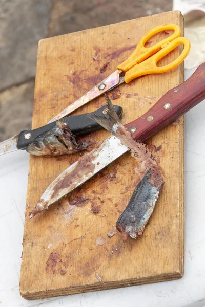 A close up view of a fishermans sardine bait on a wooden board with two knifes, yellow handle scissors that has been cut up and used to catch fish — Stock Image Close View Fishermans Sardine Bait Wooden Board Two Knifes Yellow Stock Photo