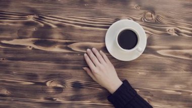 Tahta masada beyaz bir fincan kahve ve kazaklı genç bir kızın elleri. Kadınlar restoranda kahve içer. Yukarıdan bak. Sıcak bir içecekle ısıtılmış..