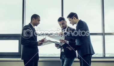 Genç takım ceketler işadamlarından geniş panoramik pencere dikilip kağıtları grafikleriyle gözden geçirme Proje tartışabiliriz. Afrikalı-Amerikalı bir işadamı ile birlikte uluslararası takım.