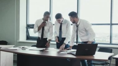 Üç işadamları, iki beyaz ve stend gömlek masada ve fırtınalı bir Afrika Milli takım proje, kağıtları çizelgeleri ile tartışın. İş takım çalışmalarını. Beyin fırtınası. Ofis panoramik pencerelere.