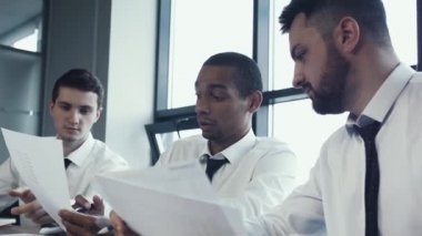 Üç işadamları, iki beyaz ve bir Afrika Ulusal, genç takım gömlek masada oturup ve projeyi, kağıtları çizelgeleri ile tartışmak. İş takım çalışmalarını. Beyin fırtınası. Ofis panoramik pencerelere.