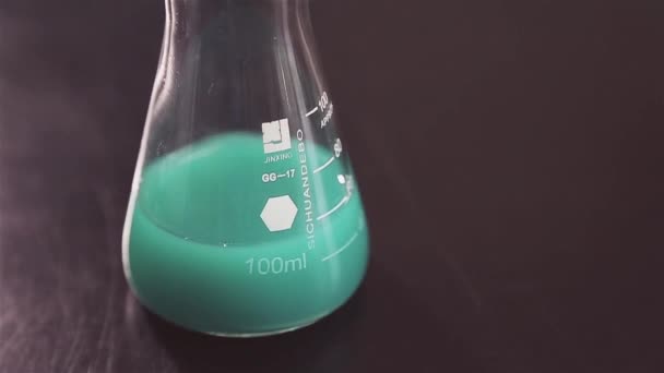 薄緑色の液体を持つフラスコは 研究室の暗いテーブルの上にあります フラスコはふたで閉じている 実験を行う 細菌用媒体 — ストック動画