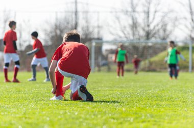 Kırmızı formalı genç futbolcu bağcıklarını yarışma arasında bağlamış.