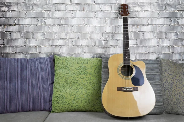 白いレンガの壁の背景の上に枕を持つソファーにアコースティック ギター — ストック写真