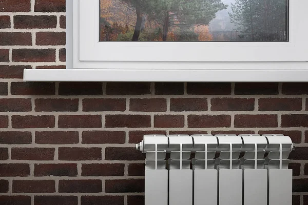 Beyaz Seksiyonel radyatör kahverengi tuğladan bir duvar bir beyaz pencere altında arka plan üzerinde Isıtma. Görüntü yazıt için boş bir alana sahip.
