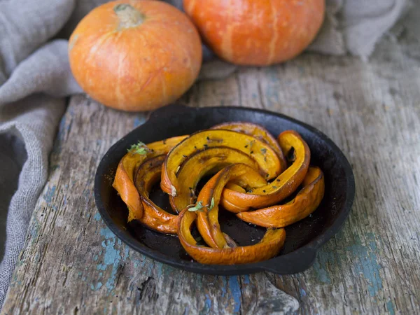 Traditional autumn dishes from pumpkin. Fried baked on grill pumpkin with spices, olive oil, herbs in a cast-iron frying pan and two orange pumpkins on an old wooden background. Close up, copy space