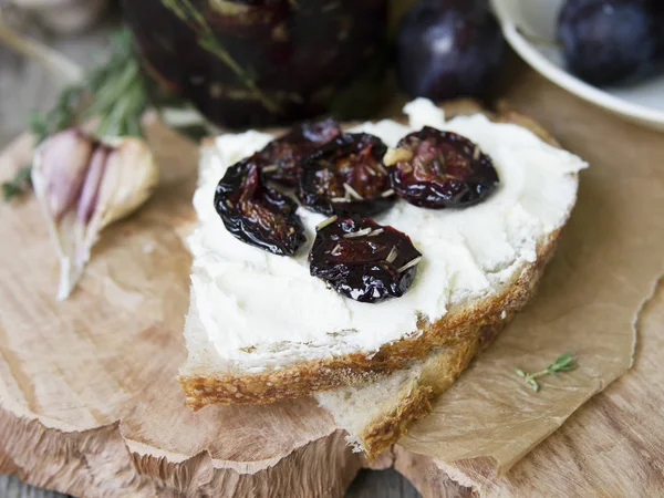 Glass jar with dried plums and fresh rosemary on wooden serving board, selective focus. Sandwich with gray bread, cream cheese and dried plums