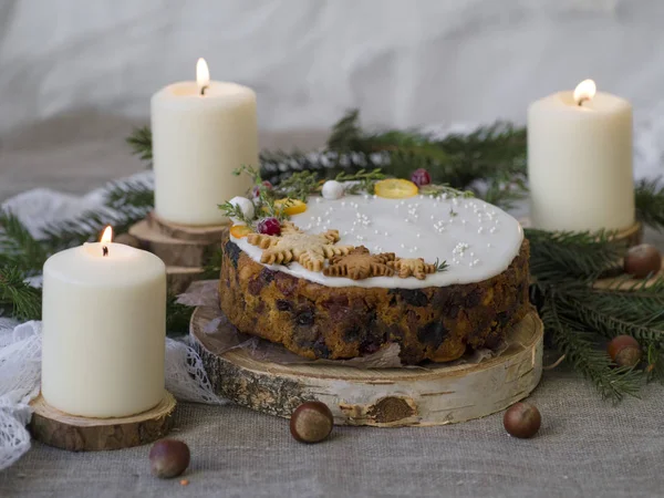 Christmas English fruitcake with candied fruit, dried fruit and nuts, decorated with white icing on a wooden background with fir branches, candles. Festive English cuisine — Stock Photo, Image
