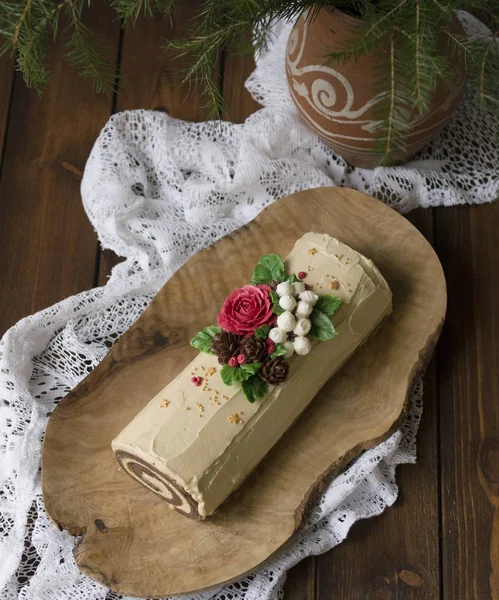 Weihnachten Biskuitrolle verziert mit cremefarbenen Blumen - Weihnachtsstern, Baumwolle, Rose, Zapfen - auf einer hölzernen Schale auf Holzgrund. Schokoladenkuchen. Weihnachts- oder Neujahrsthema — Stockfoto