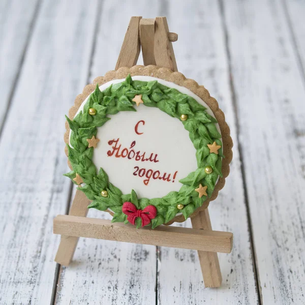 A glaze Christmas gingerbread cookie in the form of a Christmas wreath with a red bow and golden stars with inscription in Russian Happy New Year on a white wooden table. Close up, selective focus — Stock Photo, Image