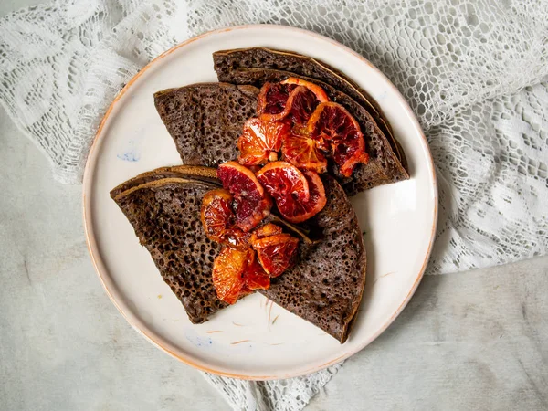 Crêpes au chocolat minces avec sauce aux oranges sanguines sur une assiette blanche sur fond gris avec du tissu en dentelle. Empilement de crêpes, blin russe, Maslenitsa. Bon petit déjeuner. Gros plan, focus sélectif — Photo