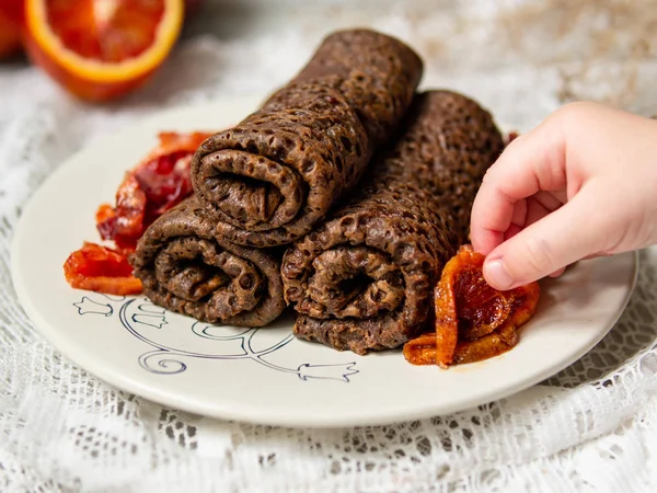 Crêpes au chocolat minces roulées dans un tube avec sauce aux oranges sanguines sur une assiette blanche avec main d'enfant. Empilement de crêpes, blin russe, Maslenitsa. Bon petit déjeuner. Concentration sélective. gros plan, espace de copie — Photo