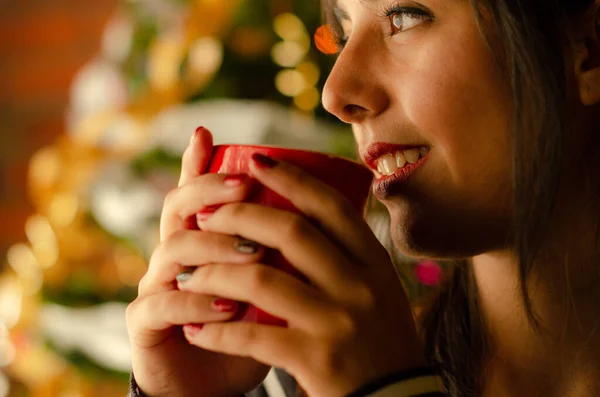 Girl drinking coffee in winter with the Christmas tree behind her while looking out the window