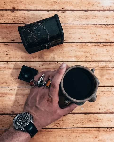 Aerial photo. Man\'s hand with multiple rings on his fingers takes his coffee cup in the shape of a head, which is placed on a wooden table