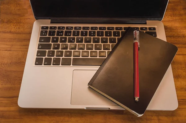 Black notebook with a pen to write work notes on the keyboard of a laptop computer, these office supplies are on a desk.