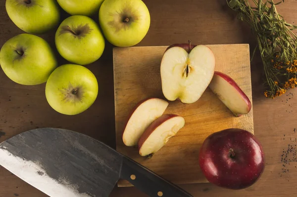Red apple cut into slices on a kitchen board and a knife, surrounded by green apples.