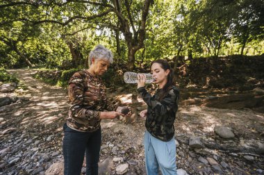 Anne ve kızı nehir kıyısında yürürken kızı plastik bir şişeden su içiyor..