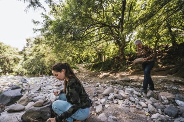 Anne ve kızı nehrin kenarında apacheta yaparken eğleniyorlar, gülümsüyorlar ve anın tadını çıkarıyorlar..