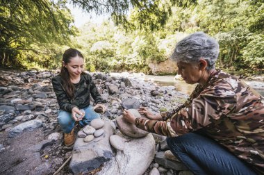 Anne ve kızı nehrin kenarında apacheta yaparken eğleniyorlar, gülümsüyorlar ve anın tadını çıkarıyorlar..