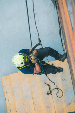 Firefighter rescuer descending a massive wall using a rappelling device in a multi-story building, wearing protective gear such as a helmet, harness, and carabiners.