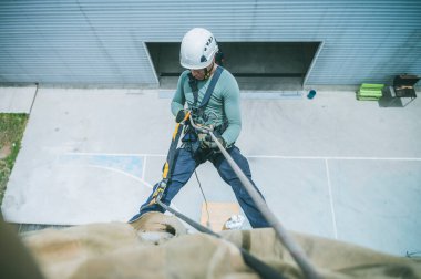 Firefighter rescuer descending a massive wall using a rappelling device in a multi-story building, wearing protective gear such as a helmet, harness, and carabiners.
