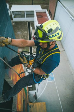 Firefighter rescuer descending a massive wall using a rappelling device in a multi-story building, wearing protective gear such as a helmet, harness, and carabiners.