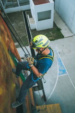 Firefighter rescuer descending a massive wall using a rappelling device in a multi-story building, wearing protective gear such as a helmet, harness, and carabiners.
