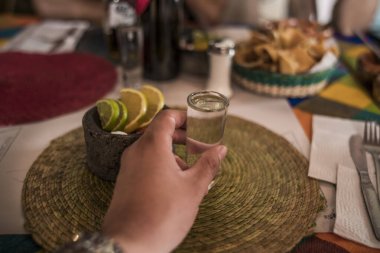 POV photo with hand and delicious crystalline mezcal in its glass caliber, accompanied by worm salt, oranges, and lemon served in a small molcajete.