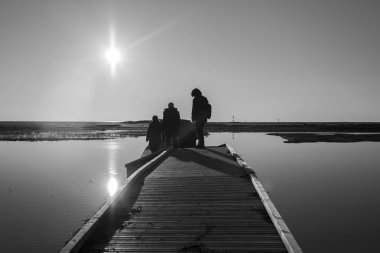 Gün batımında bir laguna üzerinde ahşap bir pontoon üzerinde yürüyen insanların arkadan aydınlatmalı siluet