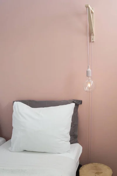 Close up on a bed with white bedding sheets and pillow, with a pastel pink wall background and design side lamp